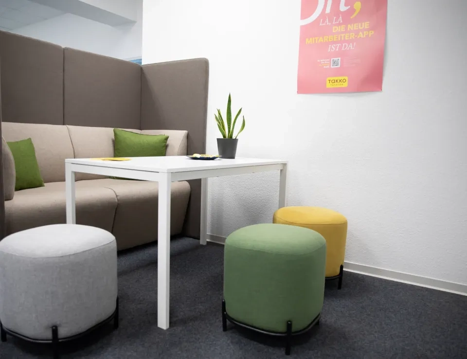 Eine moderne Bürolounge mit einem weißen Tisch, einem grauen Sofa, grünen und gelben Kissen, drei runden Hockern und einer Topfpflanze. An der weißen Wand über dem Tisch hängt ein rosa Poster.