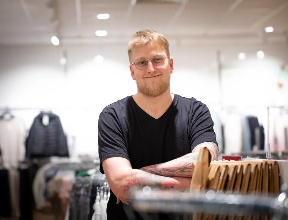 Ein lächelnder Mann mit Brille und Tattoos auf dem linken Arm steht mit verschränkten Armen in einem Bekleidungsgeschäft, umgeben von Kleiderständern. Der Hintergrund ist leicht verschwommen.