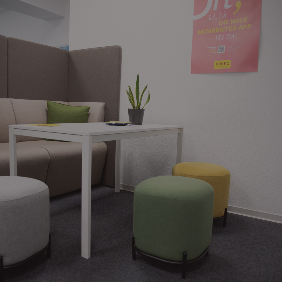 Moderne Bürolounge mit beigem Sofa, weißem Tisch, grüner Pflanze und drei runden Hockern in Grau, Grün und Gelb. Über dem gelben Hocker hängt ein rosa Poster an der Wand.
