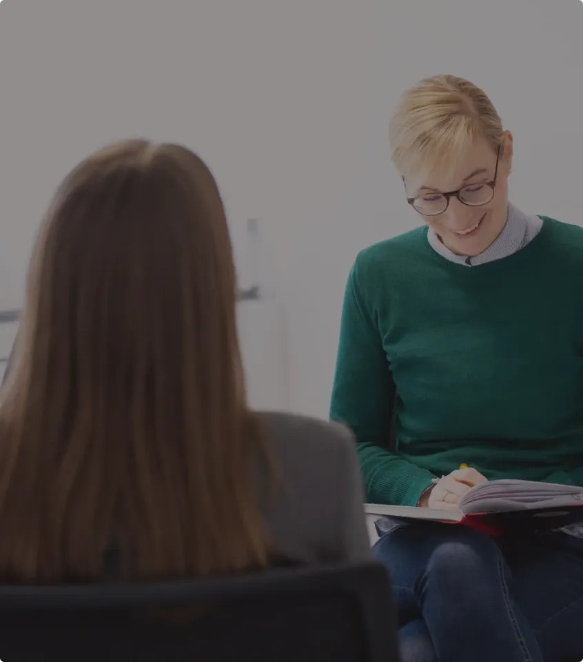 Eine Frau in einem grünen Pullover und mit Brille lächelt, während sie in einem hellen Innenraum während eines Gesprächs mit einer anderen Frau, die der Kamera den Rücken zuwendet, in ein Notizbuch schreibt.