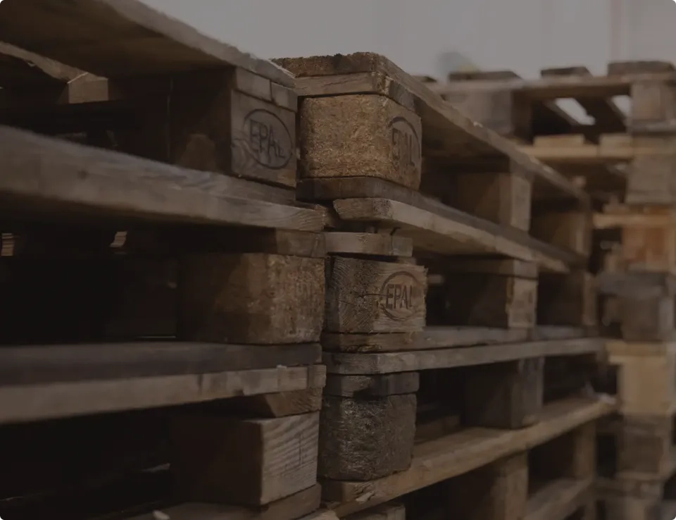 Eine Nahaufnahme gestapelter Holzpaletten, die ihre raue Textur und ihr verwittertes Aussehen in einem schwach beleuchteten Lagerbereich zeigt.
