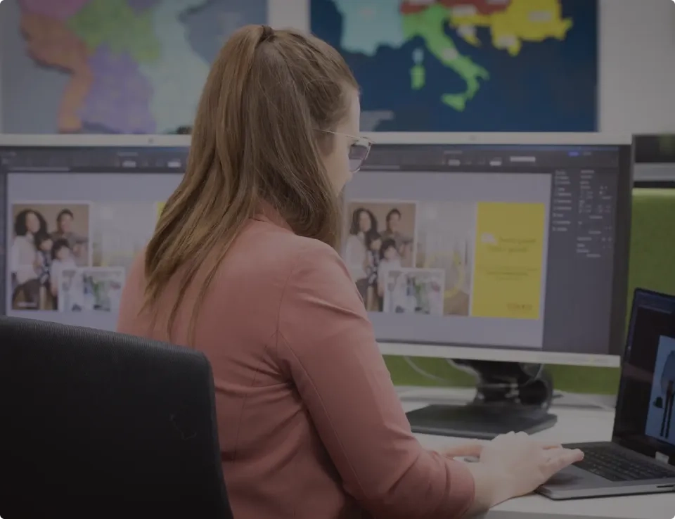 Eine Frau mit langen braunen Haaren und Brille arbeitet an einem Schreibtisch mit zwei Computerbildschirmen, auf denen Grafiksoftware und Familienfotos laufen. Im Hintergrund hängt eine bunte Landkarte an der Wand.