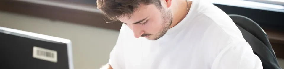 Ein junger Mann mit kurzen braunen Haaren und einem weißen T-Shirt sitzt an einem Schreibtisch und konzentriert sich auf etwas vor ihm. In der Nähe steht ein Computermonitor.