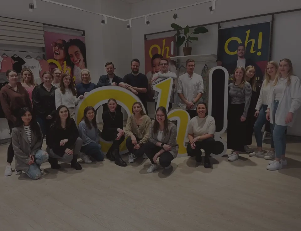 Eine Gruppe von etwa zwanzig Personen posiert gemeinsam in einem Innenbereich vor bunten Schildern mit der Aufschrift „Oh!“. Einige stehen, andere sitzen und alle lächeln in die Kamera.