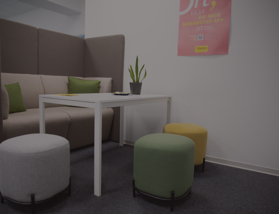 Eine moderne Bürolounge mit einem weißen Tisch, grauen und grünen Ottomanen, einem beigen Sofa mit grünen Kissen, einer Pflanze auf dem Tisch und einem rosa Poster an der Wand.