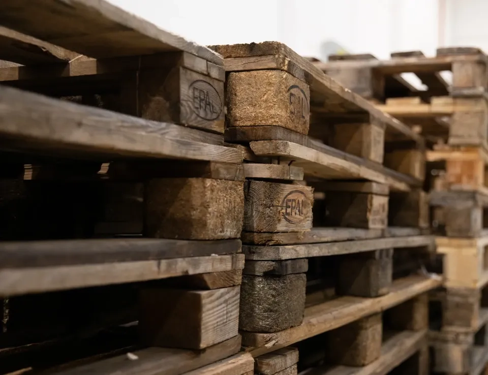 Nahaufnahme gestapelter Holzpaletten mit ihrer rauen, verwitterten Textur und Markierungen. Die Paletten sind in einem Innenbereich angeordnet.
