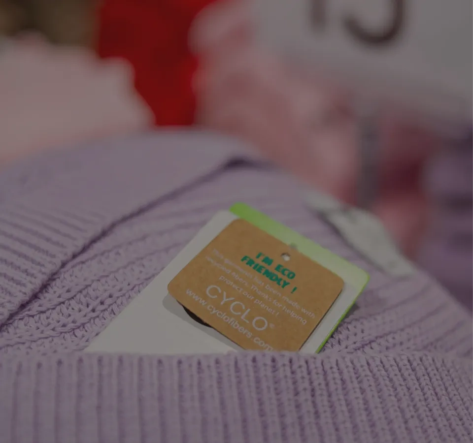 Nahaufnahme eines hellvioletten Strickkleidungsstücks mit einem braunen Papieretikett mit der Aufschrift „ECO FRIENDLY!“ und „CYCLO“, was darauf hinweist, dass der Artikel umweltfreundlich ist. Der Hintergrund ist leicht unscharf.