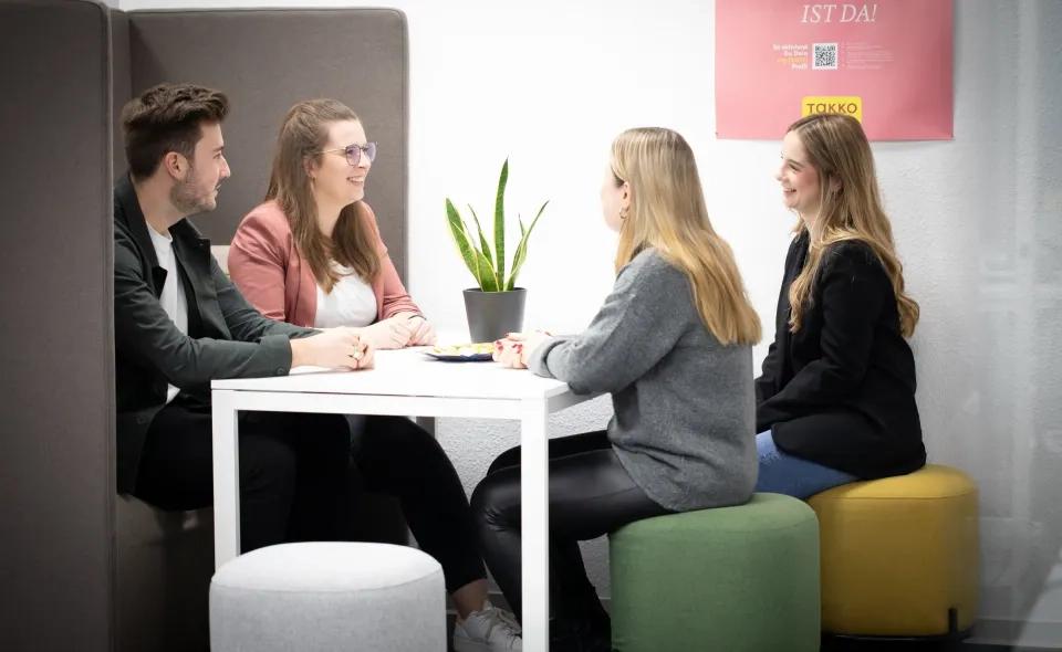 Vier Personen sitzen lächelnd und unterhalten sich um einen weißen Tisch in einem modernen Büro. Eine kleine Pflanze steht auf dem Tisch, die Personen sitzen auf bunten Hockern, hinter ihnen an der weißen Wand ein rosa Poster.