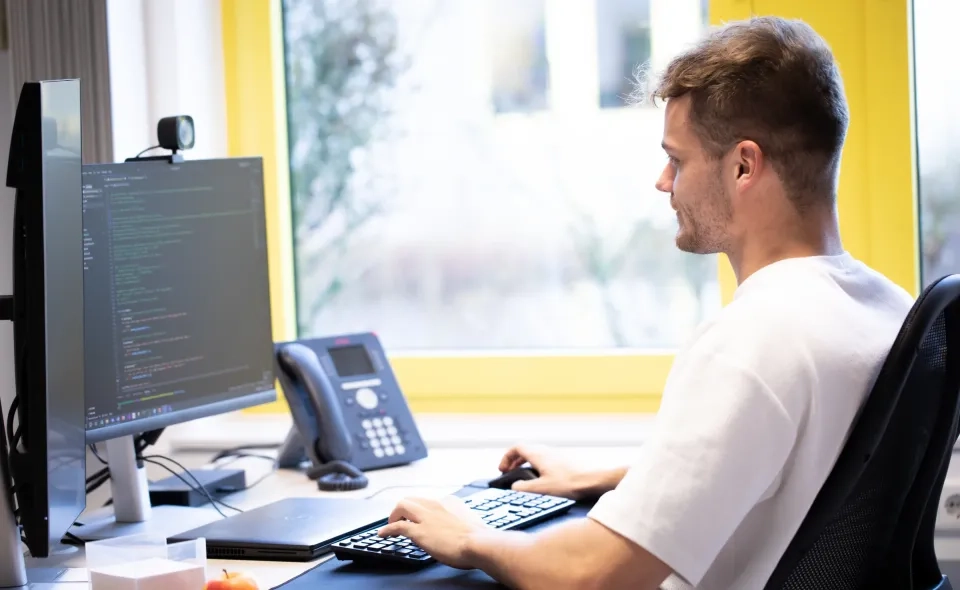 Eine Person sitzt an einem Schreibtisch vor einem Computerbildschirm, auf dem Code angezeigt wird, und tippt auf einer Tastatur. Auf dem Schreibtisch stehen ein Telefon und andere Büroartikel. Im Hintergrund ist ein leuchtend gelber Fensterrahmen zu sehen.