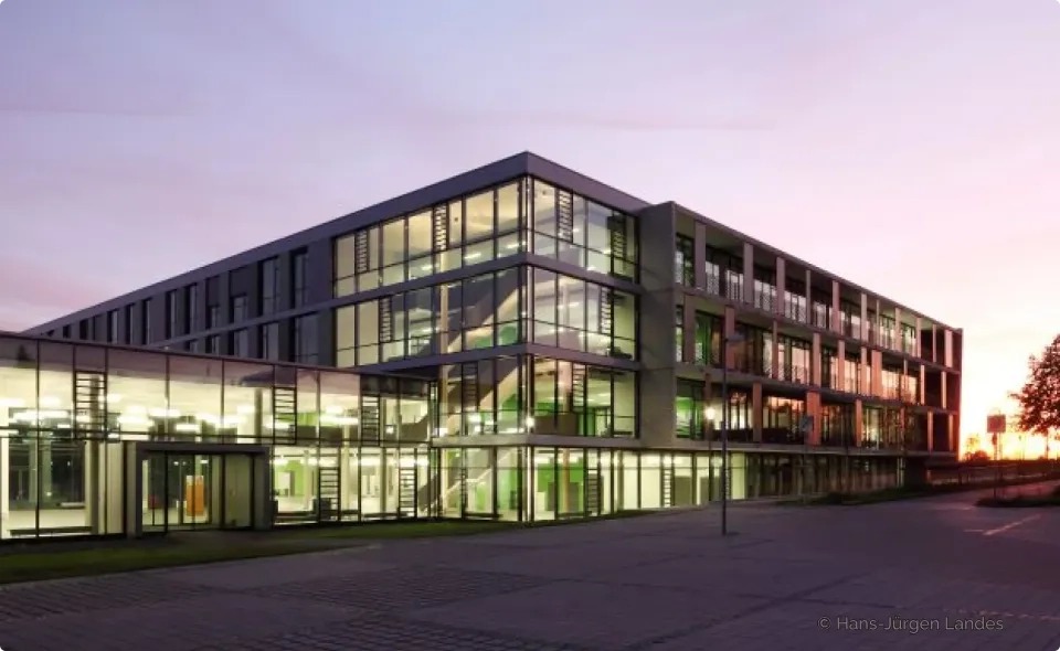 Ein modernes, mehrstöckiges Gebäude aus Glas und Stahl ist bei Sonnenuntergang zu sehen. Im Inneren ist Licht an, im Hintergrund ein violett-orangefarbener Himmel. Die Architektur zeichnet sich durch große Fenster und geometrische Linien aus.
