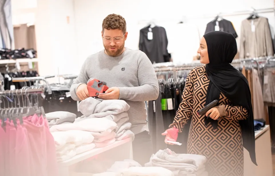 Ein Mann und eine Frau kaufen in einem Geschäft Kleidung. Der Mann betrachtet eine rote Brieftasche, während die Frau, die ein gemustertes Kleid und einen schwarzen Hijab trägt, lächelt und Kleidung hält. Sie stehen neben Kleiderständern mit gefalteten Pullovern.