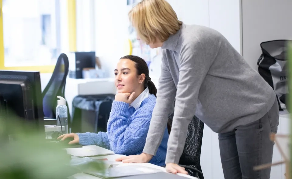 Zwei Frauen in einem Büro; eine sitzt an einem Schreibtisch und schaut auf einen Computerbildschirm, während die andere neben ihr steht, sich zu ihr beugt und redet, beide konzentriert und in die Diskussion vertieft.