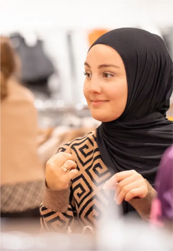 Eine Frau mit schwarzem Hijab und gemustertem Pullover sitzt drinnen, lächelt sanft und hält ihre Hände zusammen. Der Hintergrund ist verschwommen, mit undeutlichen Formen und Menschen.