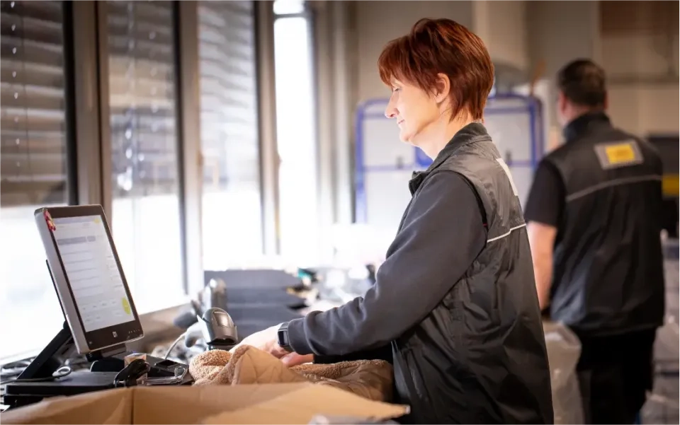 Eine Frau in einer schwarzen Weste arbeitet an einem Computerterminal in einem Lager oder einer Versandhalle. In der Nähe befinden sich ein großer Karton und ein Scanner. Im Hintergrund ist eine weitere Arbeiterin zu sehen.
