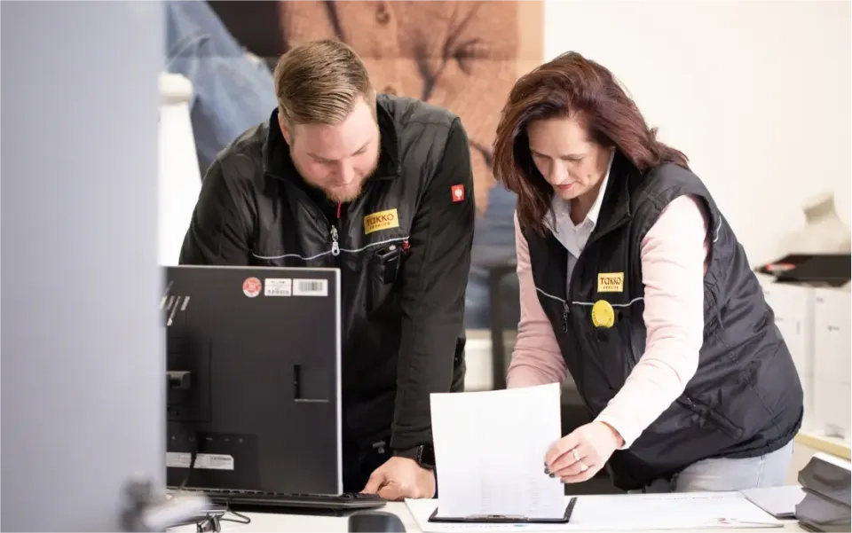 Zwei Personen in schwarzen Westen arbeiten gemeinsam an einem Schreibtisch mit Computer. Eine Person blickt auf den Monitor, die andere hält gedruckte Dokumente in der Hand und prüft sie. Beide wirken konzentriert.