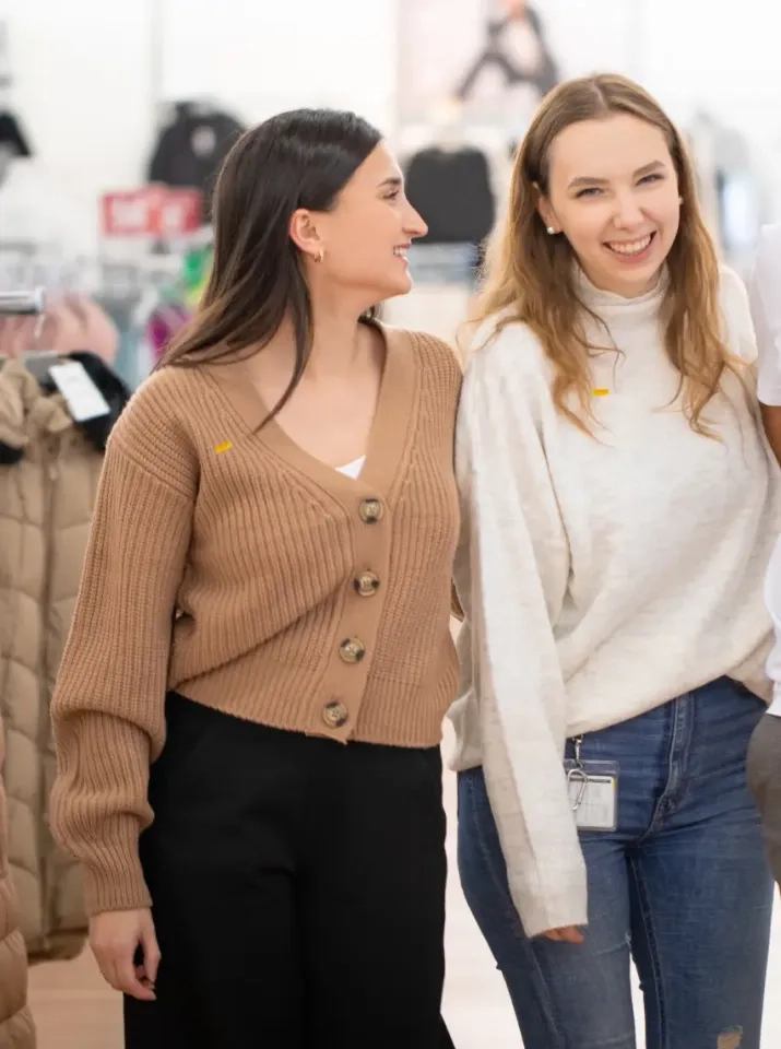 Zwei Frauen stehen nebeneinander, lächeln und wirken glücklich. Die eine trägt eine braune Strickjacke und eine schwarze Hose, die andere einen weißen Pullover und blaue Jeans. Sie scheinen sich in einem Geschäft oder einer Einkaufsumgebung zu befinden.