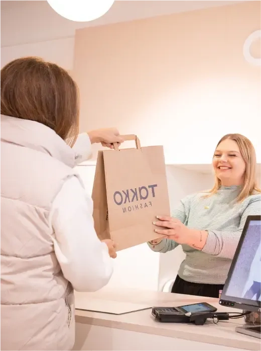 Eine lächelnde Kassiererin überreicht einem Kunden in einer hellen Jacke an einer Ladentheke, an der ein Zahlungsterminal und ein Monitor zu sehen sind, eine Einkaufstüte aus Papier mit der Aufschrift „TOKKO FASHION“.
