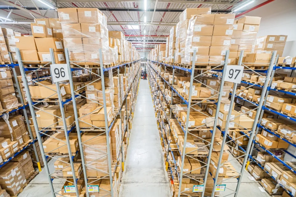 Weitwinkelansicht eines Lagerhalleninneren mit hohen Regalen voller Kartons zu beiden Seiten eines Mittelgangs. Schilder mit den Aufschriften 96 und 97 hängen an den Regalen. Helle Deckenbeleuchtung erhellt den Raum.