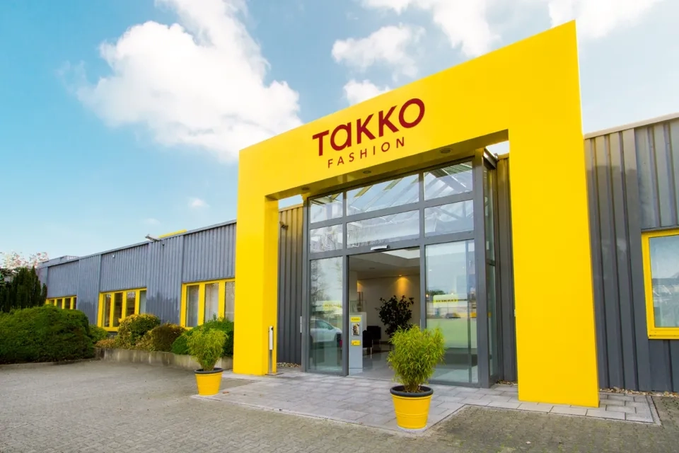 Der Eingang eines Takko Fashion Stores mit einem leuchtend gelben Rahmen, Glastüren und zwei Topfpflanzen auf beiden Seiten. Das Gebäude ist grau mit gelben Fensterrahmen, unter einem blauen Himmel mit Wolken.
