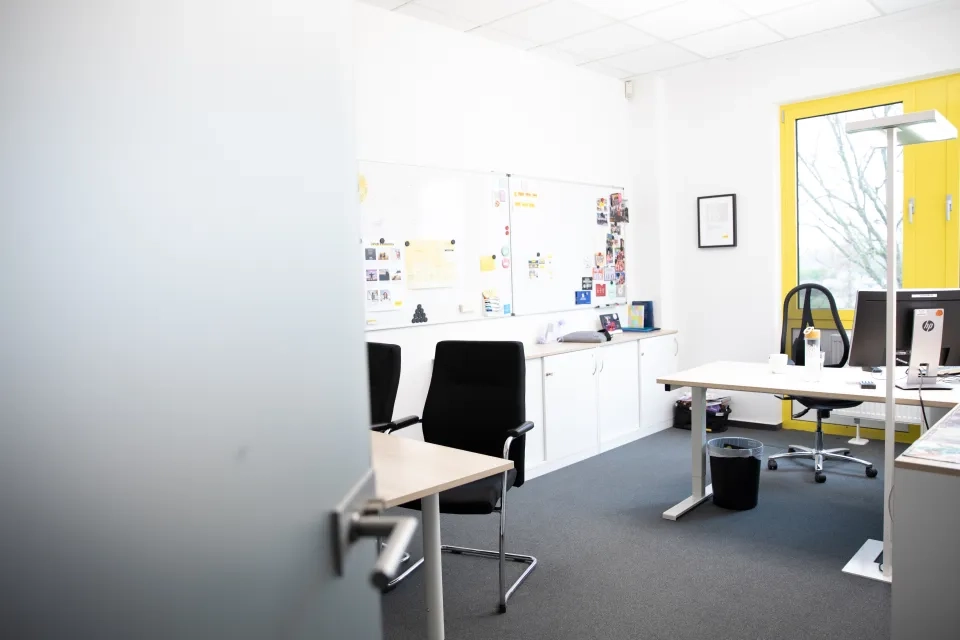 Ein helles, modernes Büro mit zwei Schreibtischen, Bürostühlen, einem Computermonitor, einem Whiteboard mit Notizen und Bildern und einem großen Fenster mit gelbem Rahmen, das natürliches Licht hereinlässt.