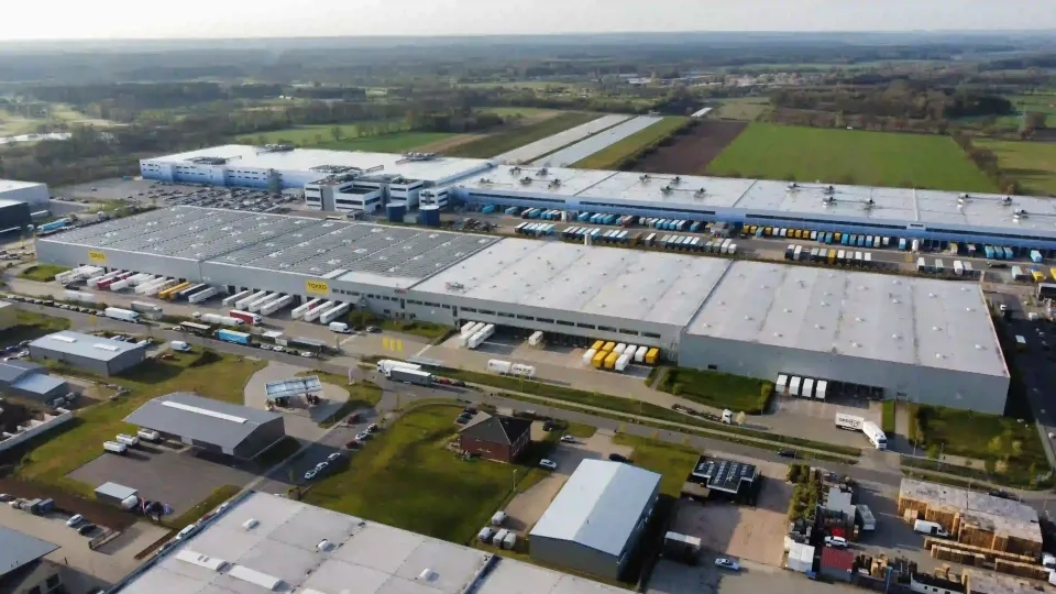Luftaufnahme eines großen Industriekomplexes mit mehreren Lagerhallen, Laderampen und zahlreichen davor geparkten LKWs. Um die Anlage herum befinden sich Felder, kleinere Gebäude und Straßen.