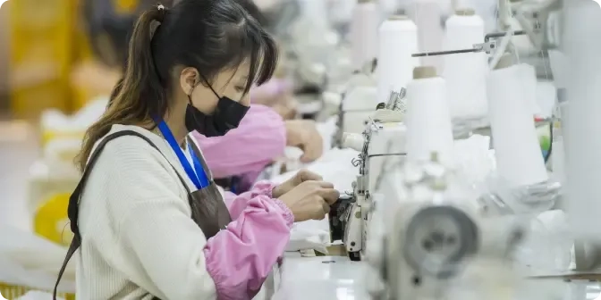Eine Frau mit Maske und Schürze arbeitet an einer Nähmaschine in einer Textilfabrik, umgeben von Fäden und anderen Arbeitern an ähnlichen Stationen.