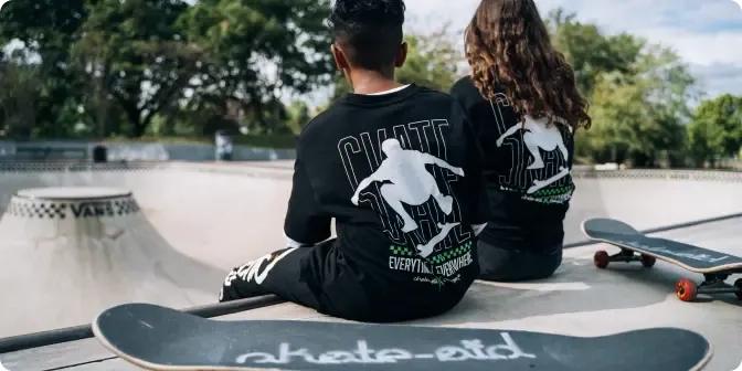 Zwei Personen sitzen mit dem Rücken zur Kamera am Rand einer Skatepark-Bowl und halten jeweils ein Skateboard in der Hand. Sie tragen passende schwarze Shirts mit Skateboard-Grafiken. Im Hintergrund sind Bäume und blauer Himmel zu sehen.