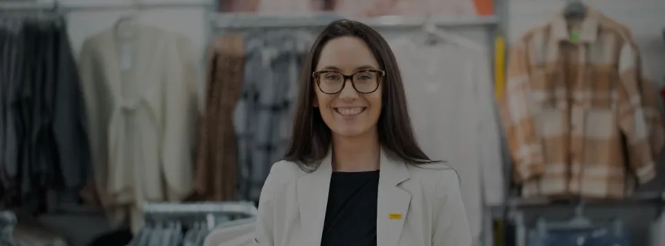 Eine Frau mit langen braunen Haaren und Brille, die einen weißen Blazer und ein schwarzes Hemd trägt, lächelt, während sie in einem Bekleidungsgeschäft steht, hinter dem sich Kleiderständer mit Kleidung befinden.