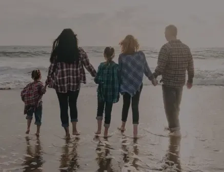 Fünf Menschen – zwei Erwachsene und drei Kinder – gehen Hand in Hand an einem Strand entlang, mit Blick aufs Meer. Sie tragen legere, karierte Hemden und Jeans, und ihre Spiegelbilder sind im nassen Sand sichtbar.