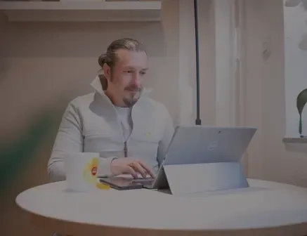 Ein Mann mit zurückgebundenen Haaren sitzt an einem runden weißen Tisch und benutzt einen Laptop. Eine Tasse und ein Smartphone liegen auf dem Tisch. Er wirkt konzentriert, in einem sanft beleuchteten Raum mit einem Fenster in der Nähe.