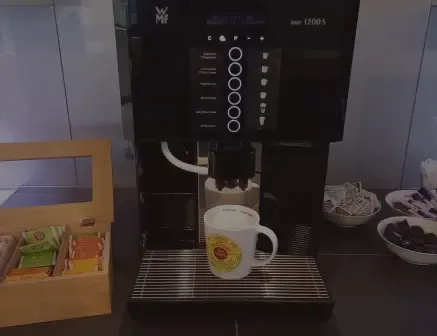 Eine Kaffeemaschine füllt Kaffee in eine weiße Tasse. Links steht eine Holzbox mit Teebeuteln, rechts stehen Schalen mit Zuckerpäckchen und Kaffeeweißer.