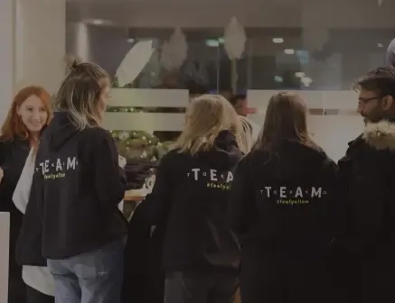 Eine Gruppe von Menschen (von hinten zu sehen) trägt passende schwarze Kapuzenpullover mit den Aufdrucken TEAM und #lovetofollow auf dem Rücken. Sie sitzen um einen Tisch im Innenbereich, eine Frau lächelt in die Kamera.
