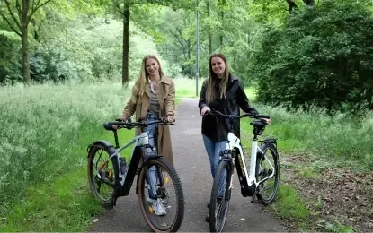 Zwei Frauen stehen mit Fahrrädern auf einem Waldweg und lächeln in die Kamera. Üppige grüne Bäume und Gras umgeben sie und schaffen eine friedliche Atmosphäre.