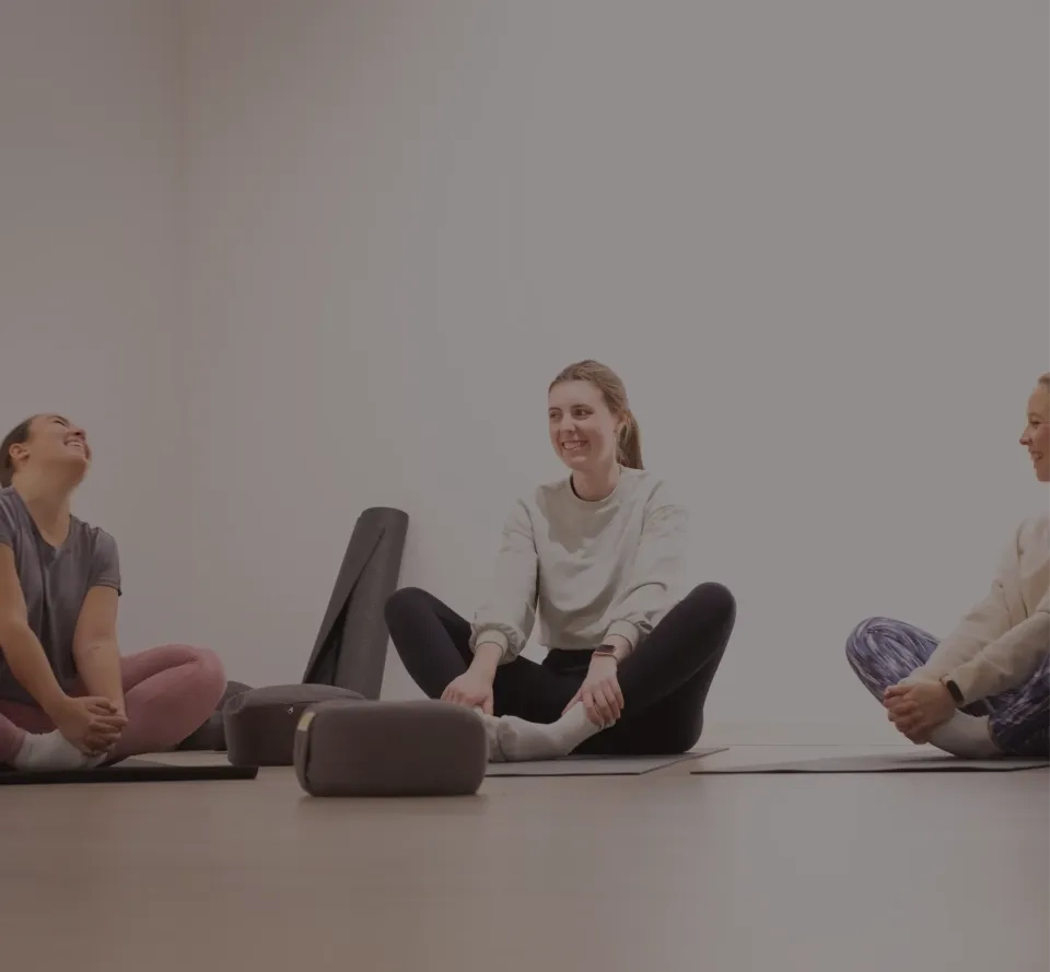 Drei Frauen sitzen lächelnd auf Yogamatten in einem hellen Raum und machen Dehnübungen. Yoga-Requisiten stehen bereit. Die Atmosphäre wirkt entspannt und freundlich.
