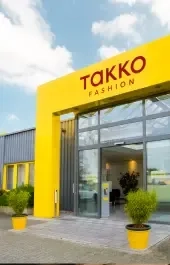 Der Eingang eines Takko Fashion-Geschäfts mit einer leuchtend gelben Fassade, großen Glastüren und Topfpflanzen auf beiden Seiten des Eingangs unter einem teilweise bewölkten Himmel.