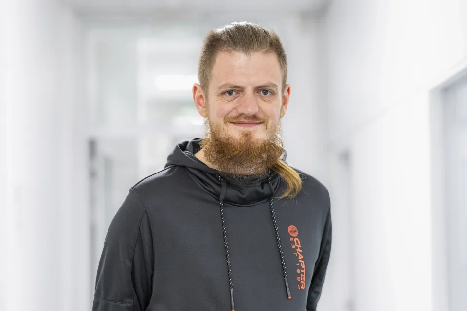 Ein Mann mit langem braunem Bart und Pferdeschwanz, der einen schwarzen Kapuzenpullover mit orangefarbener Aufschrift trägt, steht lächelnd in einem hellen, weißen Flur.