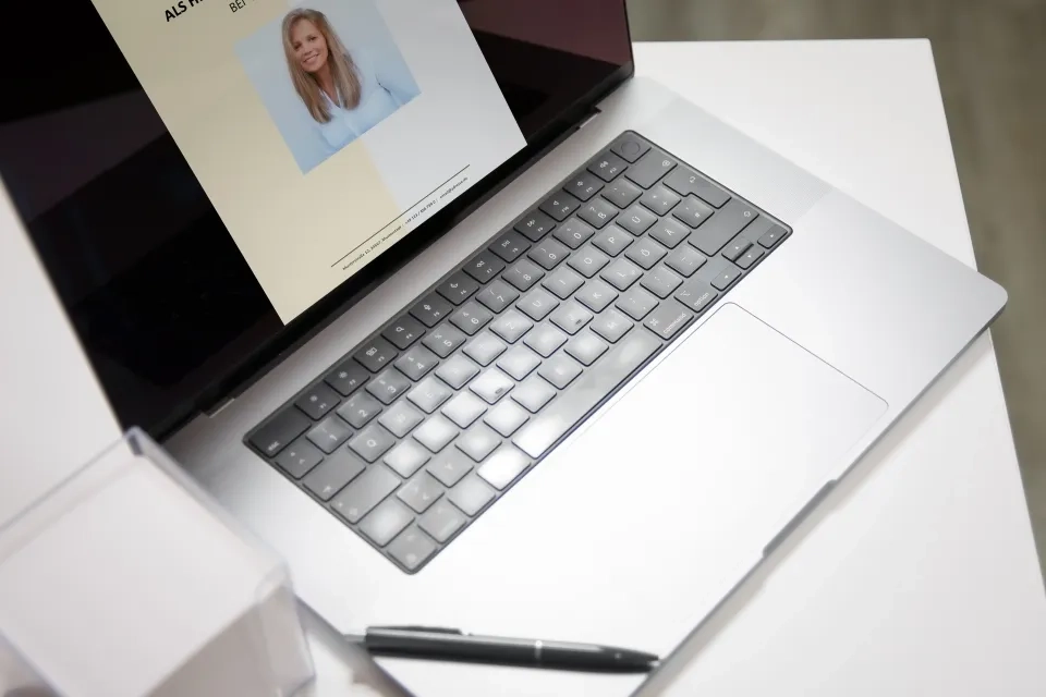 Ein silberner Laptop steht auf einem weißen Schreibtisch. Auf dem Bildschirm ist das Profilbild einer Frau zu sehen. Ein schwarzer Stift liegt neben der Tastatur. Das Bild ist hell und klar und lässt einen professionellen Arbeitsplatz vermuten.