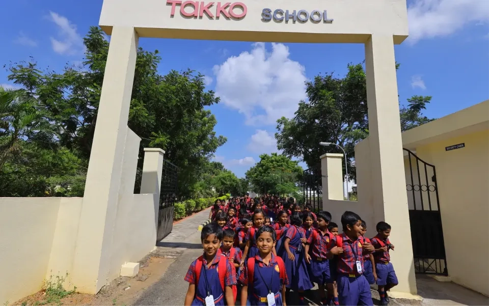 Eine große Gruppe junger Schüler in passenden Uniformen kommt an einem sonnigen Tag aus dem Eingangstor einer Schule mit der Aufschrift „TAKKO SCHOOL“, im Hintergrund Grün und blauer Himmel.