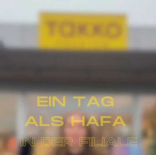 Unscharfes Bild eines Geschäfts mit einem gelben Schild mit der Aufschrift „TOKKO“. Darüber liegt in Gelb der Text „EIN TAG ALS HAFA IN DER FILIALE“.
