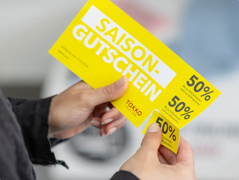 Eine Person hält einen leuchtend gelben Saison-Gutschein von Takko Fashion in der Hand, der drei Coupons mit jeweils 50 % Rabatt bietet. Der Text ist fett und weiß vor dem leuchtenden Hintergrund.