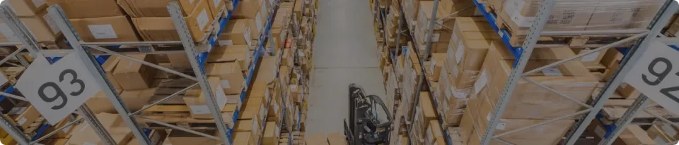 Luftaufnahme der Lagergänge mit Regalen voller Kartons. Ein Arbeiter bedient einen Gabelstapler im Mittelgang. An den Regalen sind Schilder mit den Nummern „93“ und „94“ zu sehen.