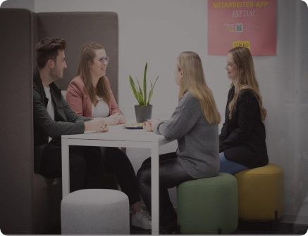 Vier junge Erwachsene sitzen in einer modernen Bürolounge um einen weißen Tisch, unterhalten sich und lächeln. Auf dem Tisch steht eine Topfpflanze, an der Wand hinter ihnen hängt ein Poster.