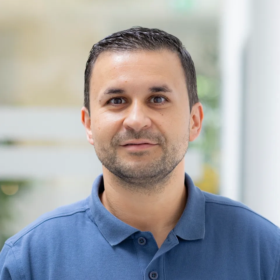 Ein Mann mit kurzen dunklen Haaren und gestutztem Bart trägt ein blaues Poloshirt und steht im Innenbereich. Er blickt mit neutralem Gesichtsausdruck direkt in die Kamera. Der Hintergrund ist leicht unscharf.