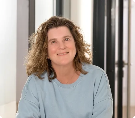 Eine Frau mit welligem braunem Haar und einem hellblauen Pullover lächelt in die Kamera, während sie in einem modernen, gut beleuchteten Raum sitzt.