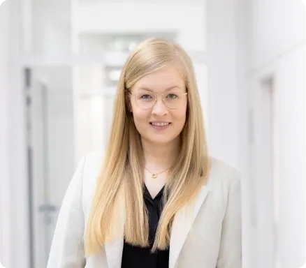 Eine Frau mit langen blonden Haaren, Brille, hellem Blazer und schwarzem Top steht in einem hellen Flur und lächelt in die Kamera.