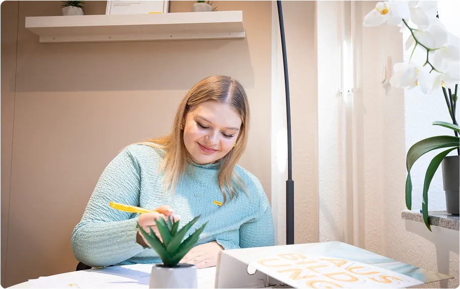 Eine junge Frau mit blonden Haaren und einem hellblauen Pullover sitzt lächelnd an einem Schreibtisch und schreibt mit einem gelben Stift. Auf dem Schreibtisch liegen Papiere, eine kleine Topfpflanze und ein Buch mit dem Titel „AUSBILDUNG“. Regale und Pflanzen schmücken den Hintergrund.