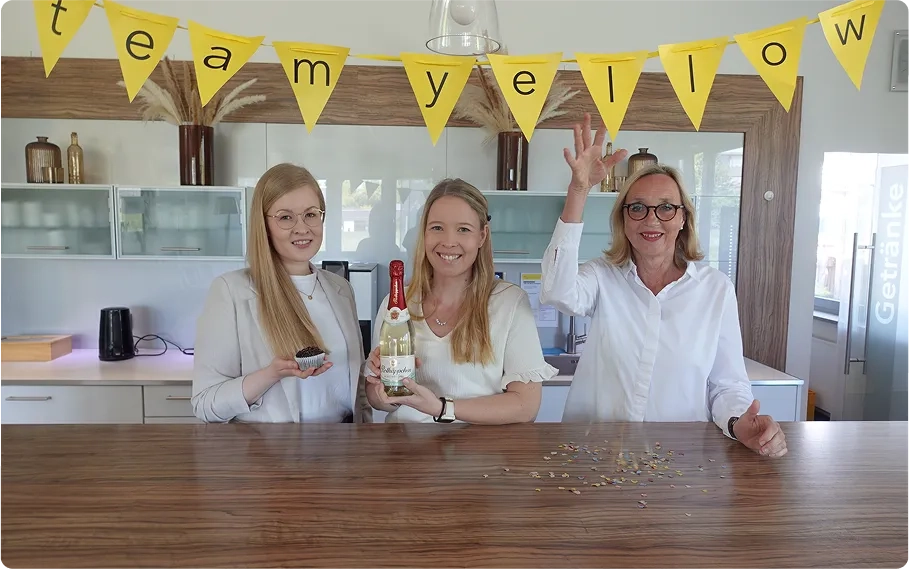 Drei Frauen stehen lächelnd hinter einem Tresen unter einem gelben Banner mit der Aufschrift „Team Gelb“. Eine hält eine Flasche, eine andere ein kleines Schälchen und die dritte gestikuliert mit der Hand über dem verstreuten Konfetti auf dem Tresen.