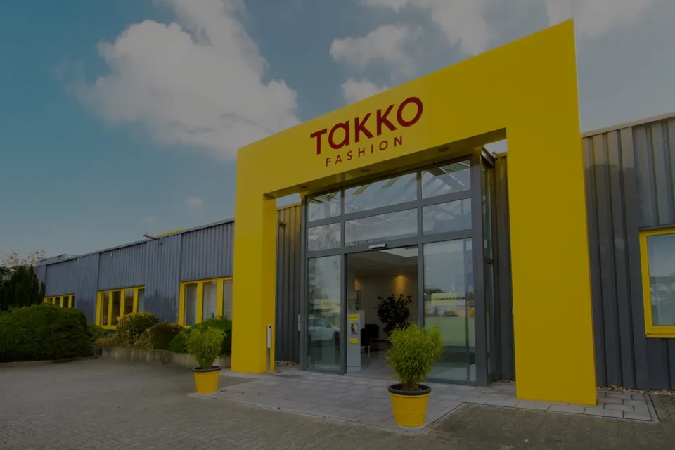 Der Eingang eines Takko Fashion-Geschäfts mit einem leuchtend gelben Rahmen, Glastüren und Topfpflanzen draußen unter einem teilweise bewölkten Himmel.