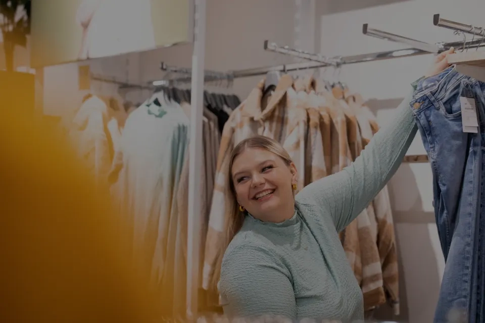Eine lächelnde Frau in einem hellblauen Oberteil hält beim Einkaufen in einem Bekleidungsgeschäft eine Bluejeans hoch, im Hintergrund sind Kleiderständer mit Kleidung und Mänteln zu sehen.