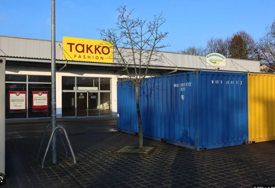 Ein Takko Fashion-Geschäft mit einem gelben Schild befindet sich hinter zwei großen Schiffscontainern, einem blauen und einem gelben, auf einer gepflasterten Fläche mit einem kahlen Baum in der Mitte.